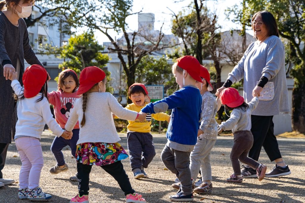 晴れた日の公園で、笑顔の保育士と赤い帽子をかぶった子どもたちが手をつなぎ、輪になって楽しく遊ぶ様子