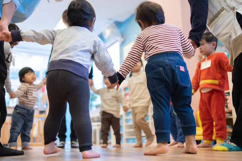 保育園で保育士と一緒に手をつなぎ、輪になって楽しく遊ぶ子どもたちの様子
